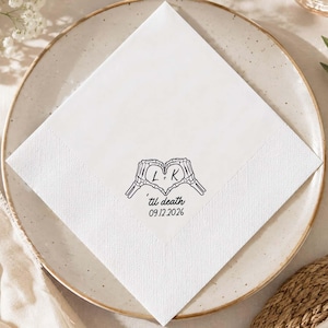 May include: A square white paper napkin featuring a black ink illustration of two skeleton hands forming a heart shape. Inside the heart are the letters L and K, with the text 'til death and the date 09.12.2026 printed below the hands.