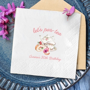 May include: White paper napkin with the text "let's par-tea" and "Aurora's 30th Birthday" in pink script. The napkin features a watercolor illustration of a teapot, teacup, and pastries. The napkin is on a blue plate.