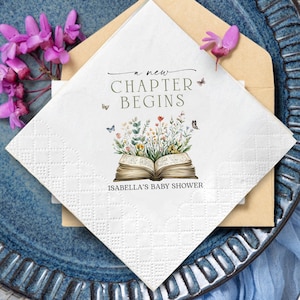 May include: White paper napkin with the text "a new CHAPTER BEGINS" and "ISABELLA'S BABY SHOWER". The napkin features an open book with floral and butterfly illustrations. The napkin is on a blue plate with purple flowers.