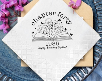 May include: White square napkin with a black floral design and text that reads "chapter forty 1985 Happy Birthday Sophia!". The napkin is placed on a blue plate with purple flowers and a tan envelope.