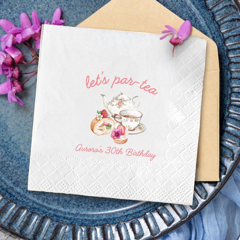 May include: White paper napkin with the text "let's par-tea" and "Aurora's 30th Birthday" in pink script. The napkin features a watercolor illustration of a teapot, teacup, and pastries. The napkin is on a blue plate.
