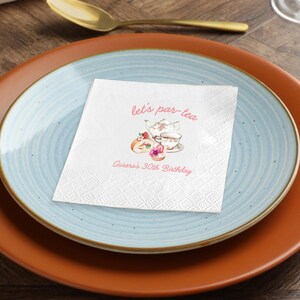 May include: A white paper napkin with a diamond pattern, featuring the text "let's par-tea" and "Aurora's 30th Birthday" in pink. The napkin has a watercolor illustration of a teapot, teacup, and pastries. The napkin is on a blue plate, which is on an orange plate.