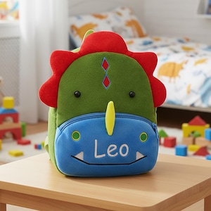 Könnte beinhalten: Ein grüner, roter und blauer Rucksack in Dinosaurierform mit dem Namen "Leo" auf der Vorderseite. Der Rucksack hat einen Reißverschluss und ein gelbes Horn. Der Rucksack steht auf einem Holztisch.