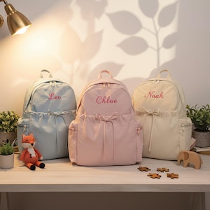 Op de afbeelding: Drie gepersonaliseerde rugzakken in pastelkleuren: lichtblauw met "Leo", roze met "Chloe" en beige met "Noah" geborduurd. Elke rugzak heeft een voorvak, zijvakken en een trekkoordsluiting. Een vosspeelgoed en houten puzzelstukjes liggen op de tafel.