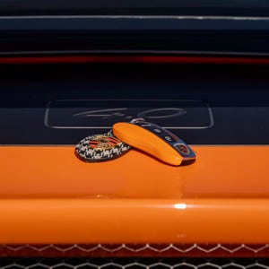 May include: An orange car key and a matching keychain rest on the hood of a car. The key is orange with black accents and the keychain has a black and white pattern with a logo. The car hood is orange and black.