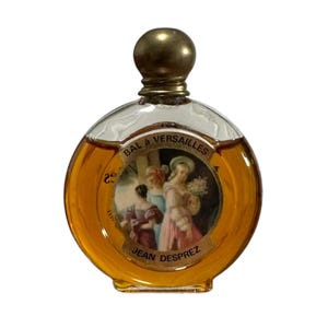 May include: A vintage perfume bottle with a round, clear glass body and a gold-colored cap. The bottle contains amber-colored liquid and features a label with the text "BAL à VERSAILLES" and "JEAN DESPREZ". The label depicts a scene with figures in period clothing.