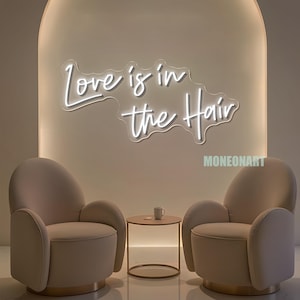 Pu&ograve; includere: Un'insegna al neon con la frase "Love is in the Hair" illumina una stanza dai toni neutri. Due poltrone beige si fronteggiano, con un tavolino rotondo e una tazza tra di loro. La stanza ha un design moderno e minimalista.