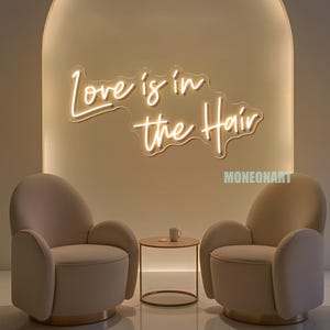 Pu&ograve; includere: Una scena d'interni moderna con due poltrone beige, un tavolino rotondo e un'insegna al neon che recita "Love is in the Hair". L'insegna &egrave; di un colore caldo e invitante, e le sedie creano un'accogliente zona salotto.