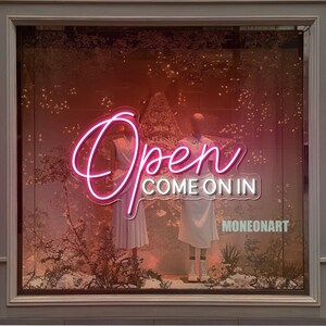 May include: A storefront window display featuring a neon sign that reads "Open COME ON IN" in pink cursive. Two mannequins in white outfits are visible behind the sign. The background has a warm, reddish-brown hue with floral and plant elements.