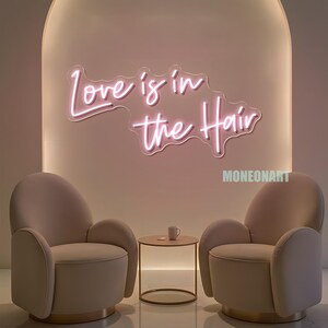 Pu&ograve; includere: Un'insegna al neon con la scritta "Love is in the Hair" in corsivo, montata su una parete rosa chiaro. Due poltrone beige si fronteggiano, con un tavolino rotondo e una tazza tra di loro. La parola "MONEONART" si trova nell'angolo in basso a destra.