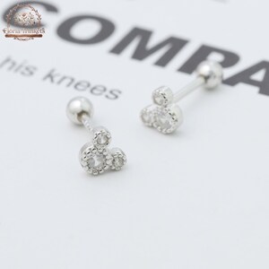 May include: A pair of silver-toned barbell earrings featuring a cluster of three small, clear, round-cut stones arranged in a triangular pattern. Each earring has a smooth, spherical backing and a metallic finish, designed for piercing.