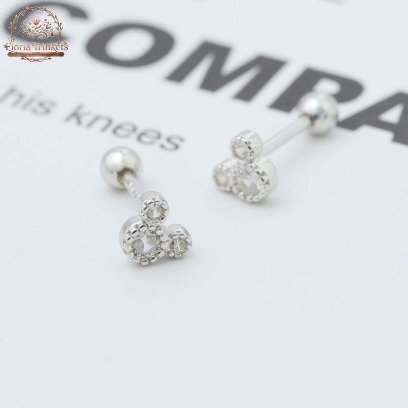 May include: A pair of silver-toned barbell earrings featuring a cluster of three small, clear, round-cut stones arranged in a triangular pattern. Each earring has a smooth, spherical backing and a metallic finish, designed for piercing.