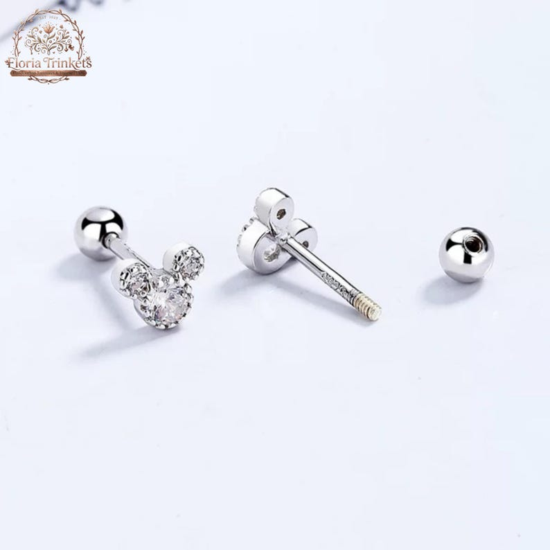 May include: Silver-toned stud earrings featuring a three-circle silhouette resembling a mouse head, accented with clear stones. The earrings include a threaded post and a spherical screw-back closure, with the Floria Trinkets logo visible.