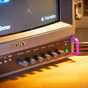 May include: A vintage Sony monitor with a silver and gray finish. The screen displays system configuration options. The monitor has several control knobs and a power button. A colorful cable and a black device are visible on the right.
