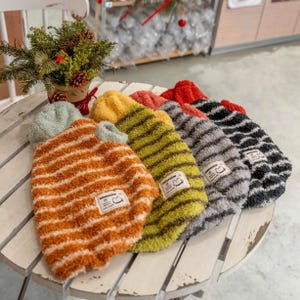 May include: A collection of striped pet sweaters in various colors, including orange, yellow, red, gray, and black. Each sweater has a small label with a cat paw print. The sweaters are arranged on a white, weathered wooden surface.