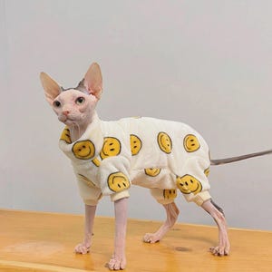 Puede incluir: Un gato Sphynx con un mono crema con patrones de caras sonrientes amarillas. El gato tiene la piel rosada y orejas grandes. El mono cubre el cuerpo y las patas del gato, con puños en los tobillos y muñecas.