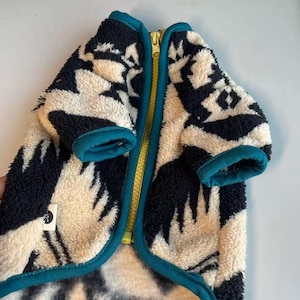 Op de afbeelding: Een hondenjas met een crèmekleurig en marineblauw geometrisch patroon. De jas heeft een gele rits, teal afwerking en manchetten. De jas is gemaakt van een zacht, pluizig materiaal.