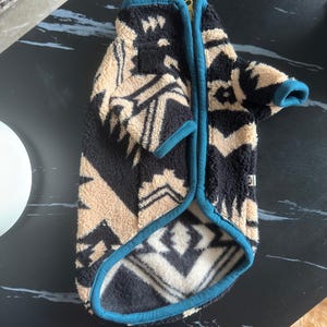 Puede incluir: Una chaqueta de forro polar para mascota con un patrón geométrico en negro y beige y ribetes en color verde azulado. La prenda incluye un cierre de cremallera, una pequeña etiqueta con la inscripción XL en el lateral y mangas cortas.