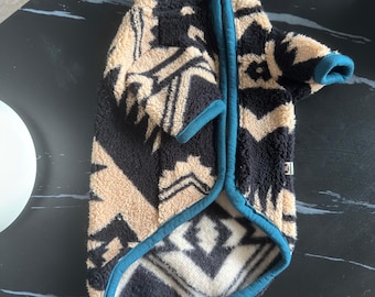 Puede incluir: Una chaqueta de forro polar para mascota con un patrón geométrico en negro y beige y ribetes en color verde azulado. La prenda incluye un cierre de cremallera, una pequeña etiqueta con la inscripción XL en el lateral y mangas cortas.