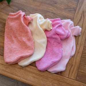 Könnte beinhalten: Vier kleine Haustierpullover in Rosa- und Cremetönen sind auf einer Holzoberfläche angeordnet. Die Pullover haben gerippte Bündchen und Ausschnitte. Die Farben reichen von einem blassen Pfirsich bis zu einem leuchtenden Rosa.