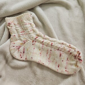 Könnte beinhalten: Eine handgestrickte Socke in Creme mit Zopfmuster und roten und gelben Sprenkeln. Die Socke liegt auf einer weichen, hellfarbigen Decke. Der Bund und die Oberseite des Fußes weisen das Zopfmuster auf.