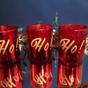 May include: Three red glass candle holders with gold glitter and the word "Ho!" written in gold script.