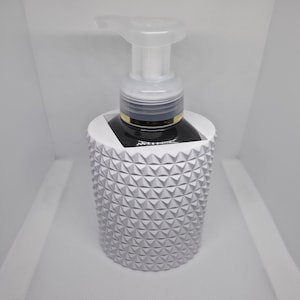 May include: A white, textured soap dispenser with a geometric pattern. The dispenser has a clear pump and a dark liquid inside. The dispenser is designed to sit on a counter or sink.