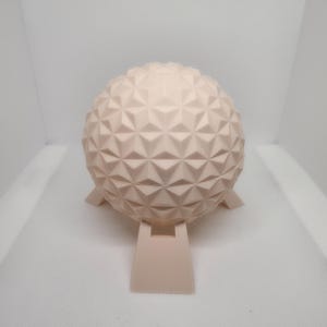 May include: A light beige, spherical decorative object with a geometric pattern. The sphere sits on a matching stand with three small feet and a rectangular base. The object has a textured surface.