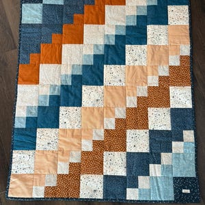 May include: A patchwork quilt with a geometric design. The quilt features squares in shades of blue, orange, peach, and white. Some squares have a dotted or patterned design. The quilt is bordered with a dark blue fabric.