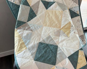 May include: A handmade quilt with a patchwork design. The quilt features geometric shapes in white, teal, yellow, and gray fabrics. The quilt is bordered with a teal fabric and is draped over a chair.
