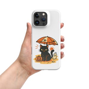 May include: White phone case featuring a Halloween-themed illustration. The design includes a black cat under an orange umbrella with bats, a jack-o'-lantern, a ghost, and a sun. The case is held in a hand.