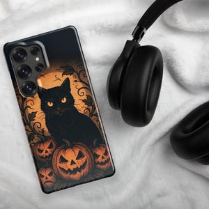 Puede incluir: Funda de teléfono negra con diseño de Halloween. Muestra un gato negro con ojos naranjas, frente a calabazas talladas. El fondo es naranja con enredaderas oscuras. Se ven auriculares negros en el fondo.