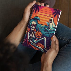 May include: Spiral-bound notebook featuring a skeleton in sunglasses relaxing on a beach with a pumpkin drink, a ghost, and a sunset. The cover art is colorful with palm trees and waves, perfect for Halloween or spooky season.