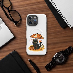 May include: White phone case featuring a Halloween-themed illustration. The design includes a black cat under an orange and white umbrella, a jack-o'-lantern, and a ghost. The case is surrounded by a wooden surface, a watch, and other office supplies.