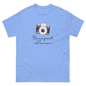 May include: Light blue t-shirt featuring a black and white camera graphic. The camera design is centered on the shirt and includes the text "Barefoot Leman" in a cursive font below the camera.