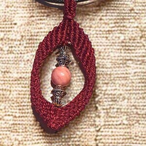 Op de afbeelding: Een bordeauxrode macramé hangerketting met een roze edelsteen en zilverkleurige accenten. De hanger hangt aan een zwart koord. De tekst "RHODONITE MACRAMÉ LIE DE VIN" staat onderaan de afbeelding.