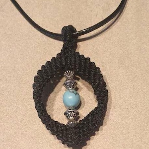 May include: Black macrame pendant necklace with a turquoise howlite bead and silver-colored accents. The pendant is an oval shape. The necklace is on a black cord.