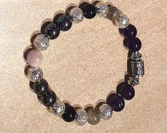 May include: A beaded bracelet featuring a mix of black, purple, silver, and light pink beads. The bracelet includes faceted silver beads and a decorative silver clasp. The beads are strung on an elastic band.