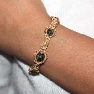 Peut inclure: Un bracelet en macramé fait à la main avec un cordon beige et des perles de pierres précieuses vert foncé. Le bracelet est porté sur un poignet, mettant en valeur le nouage complexe et le contraste entre le cordon et les perles. C'est un bijou.