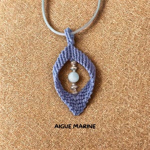 Op de afbeelding: Een handgemaakte blauwe macramé hangerketting met een lichtblauwe edelsteen kraal en zilveren accenten. De hanger heeft een druppelvorm en hangt aan een lichtblauw koord. De tekst "AIGUE MARINE" staat onderaan.