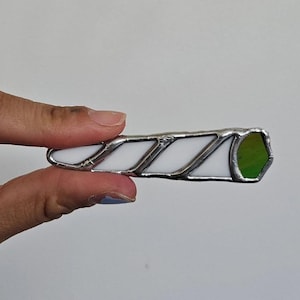 May include: A stained glass art piece in the shape of a joint. The piece is white with silver solder and a green tip. The object is held in a hand against a white background.