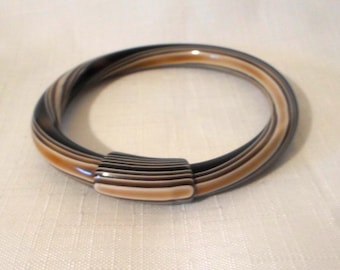 May include: A circular bangle bracelet featuring a smooth, polished finish with alternating horizontal bands of dark brown, tan, and cream colors. The bracelet includes a rectangular, ribbed connector piece that joins the circular band.
