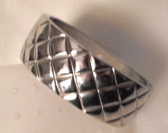 May include: A wide, polished silver-tone bangle bracelet featuring a raised, three-dimensional quilted diamond pattern across its entire outer surface. The metal has a highly reflective, smooth finish that catches the light, creating distinct highlights and shadows within each individual diamond-shaped section.