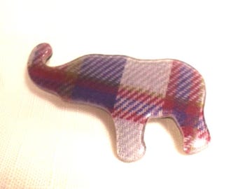 May include: An elephant-shaped brooch featuring a multi-colored plaid pattern in shades of purple, red, blue, and white. The surface has a smooth, glossy finish, and the design depicts the animal in profile with its trunk curved upward.