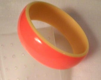 May include: A thick, rounded bangle bracelet featuring a vibrant, glossy coral-orange exterior surface and a smooth, contrasting pale cream-colored interior lining, showcasing a simple, solid-colored design with a polished finish.