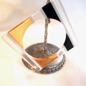 May include: A square glass coaster with a geometric design featuring clear, black, and orange sections, held upright by a silver-toned metal stand with a textured, leaf-patterned base and a slender, branch-like vertical support.