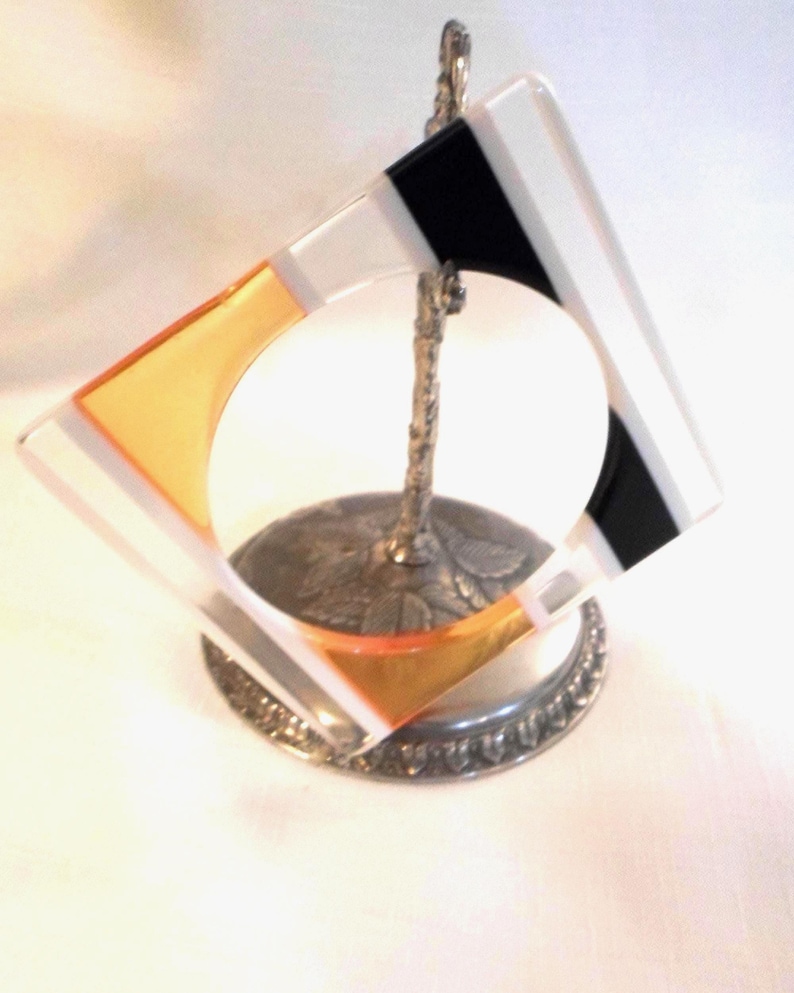 May include: A square glass coaster with a geometric design featuring clear, black, and orange sections, held upright by a silver-toned metal stand with a textured, leaf-patterned base and a slender, branch-like vertical support.