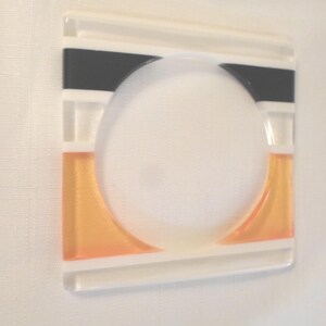 May include: A square glass coaster featuring a central circular cutout. The design includes horizontal bands of opaque black at the top and translucent orange at the bottom, separated by clear glass sections with white linear accents.