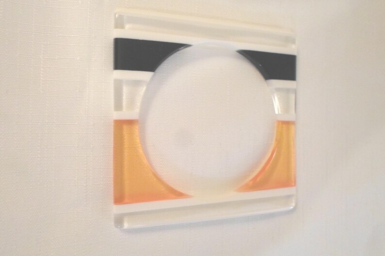 May include: A square glass coaster featuring a central circular cutout. The design includes horizontal bands of opaque black at the top and translucent orange at the bottom, separated by clear glass sections with white linear accents.