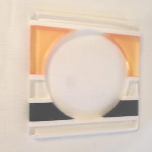 May include: A square white plastic light switch cover plate featuring a central circular cutout. The design includes a horizontal orange rectangular accent at the top and a horizontal black rectangular accent at the bottom of the plate.